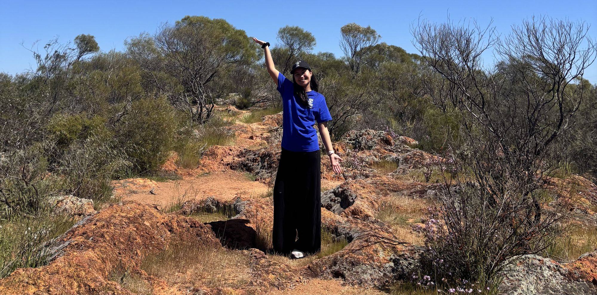 Taleena standing among red-dirt scrub with arms open under a wide Western Australian sky